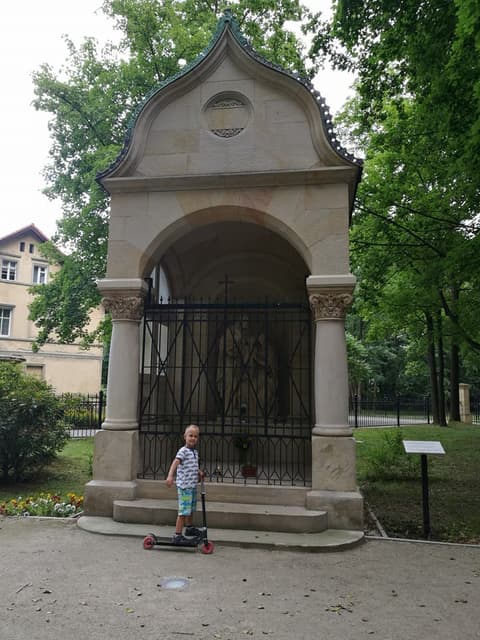Leon z hulajnogą przed zabytkową kapliczką w parku